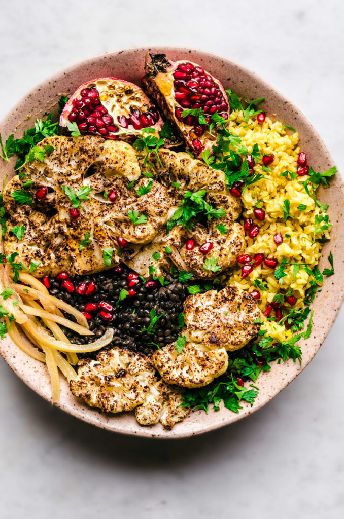 Roasted cauliflower steaks in a large bowl with preserved lemon, pomegranate, saffron rice, and black lentils.