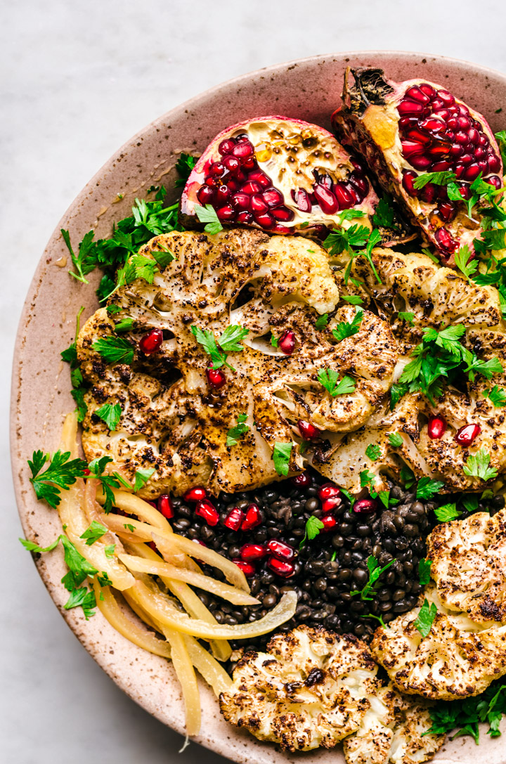 Roasted cauliflower steaks in a large bowl with preserved lemon, pomegranate, and black lentils.