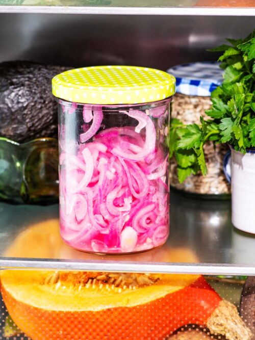 A jar of pink pickled onions in the fridge with various vegetables and herbs around.