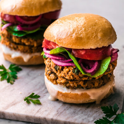 Double stacked sweet potato burger with greens, pickled onions, and tomatoes on a bun.