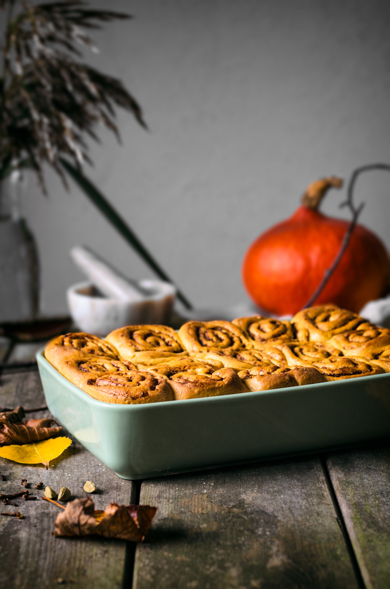 Pumpkin cinnamon rolls in baking dish with pumpkin and dishes behind the rolls.