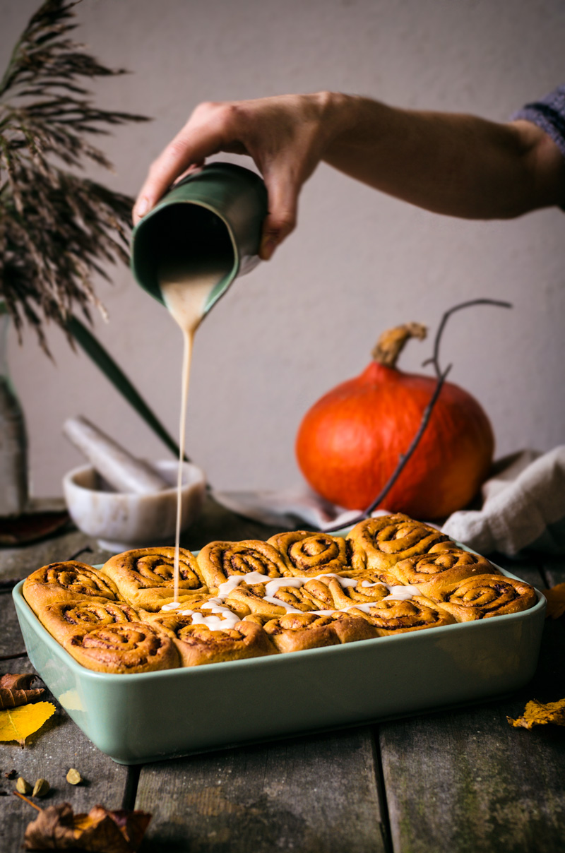 Pumpkin cinnamon rolls in baking dish with glaze being poured from small jug.