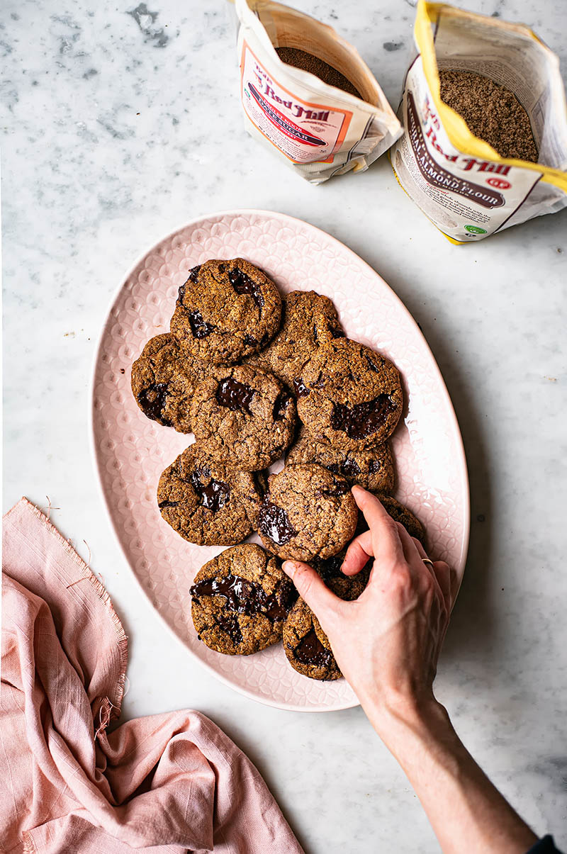 A platter of almond flour chocolate chunk cookies with branded packages in the corner and a hand reaching in.