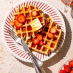 Two waffles on a plate with maple syrup and sliced strawberries.