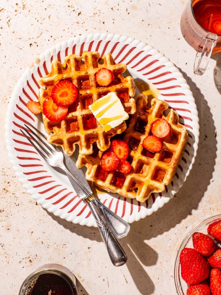 Two waffles on a plate with maple syrup and sliced strawberries.