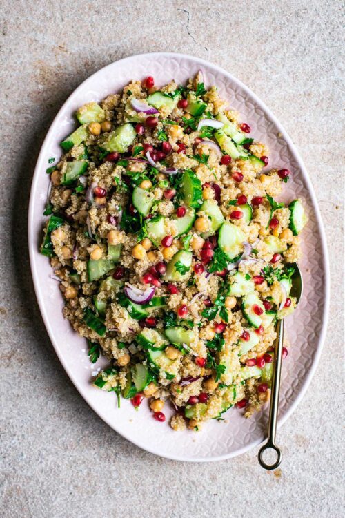 Top view of quinoa salad with chickpeas, vegetables, and pomegranate on a pink platter.