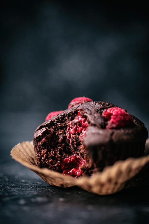 Chocolate raspberry muffin, close up in a paper liner, with a bite taken out to show interior texture.
