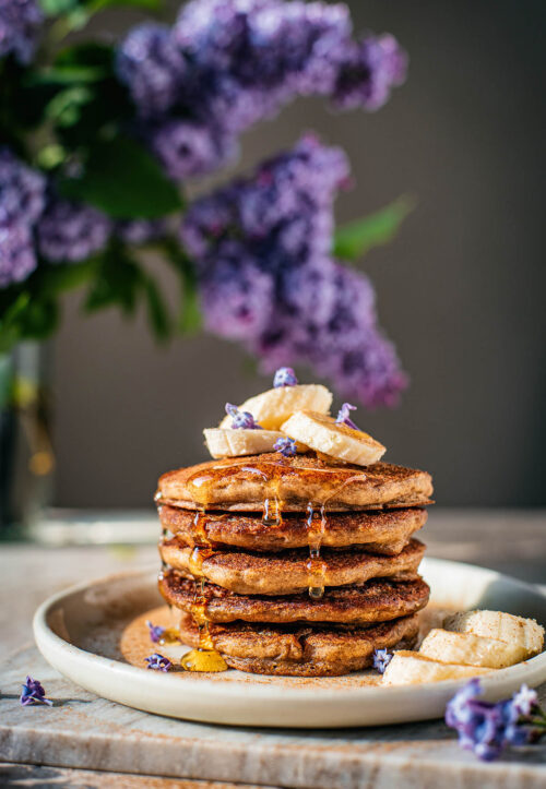 Stack of pancakes topped with bananas and lilac blossoms.