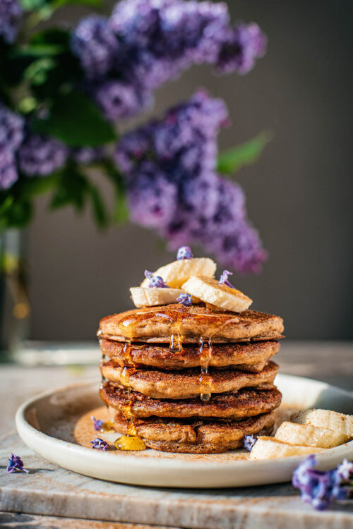 Stack of pancakes topped with bananas and lilac blossoms.