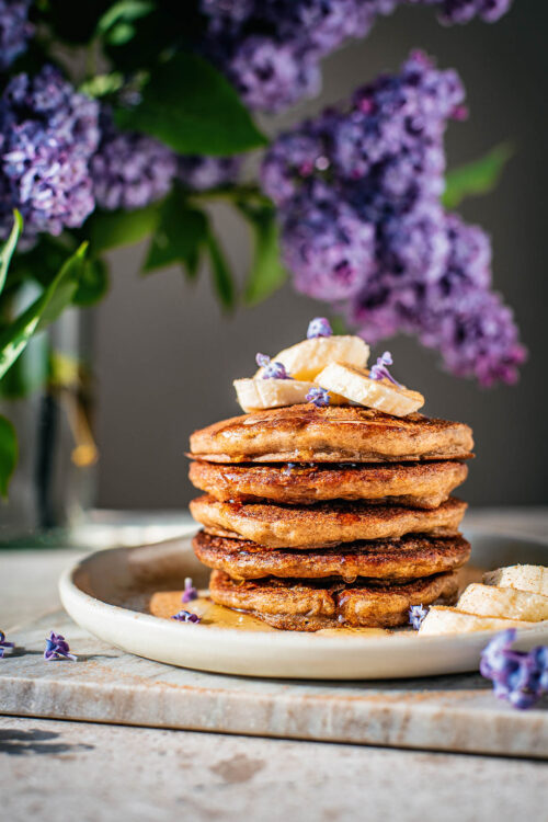 Stack of gluten free banana pancakes topped with bananas and lilac blossoms.