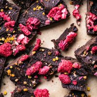 Close up of broken pieces of raw chocolate with dried raspberries, bee pollen, and dried flowers on parchment.
