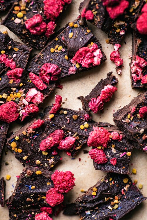 Close up of broken pieces of raw chocolate with dried raspberries, bee pollen, and dried flowers on parchment.