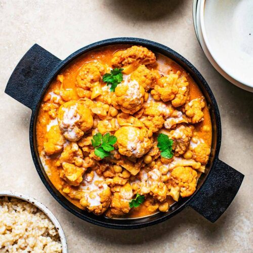 Cauliflower curry with cilantro in a cast iron pan, surrounded by brown rice in a bowl, cilantro, and empty bowls for serving.
