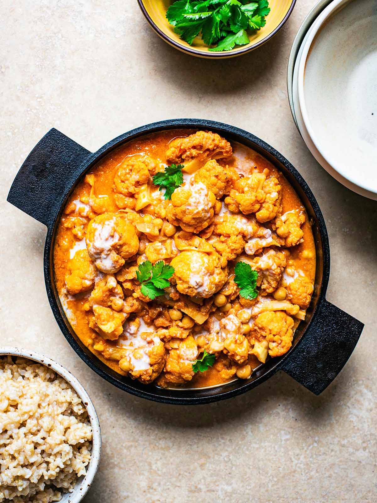 Cauliflower curry with cilantro in a cast iron pan, surrounded by brown rice in a bowl, cilantro, and empty bowls for serving.