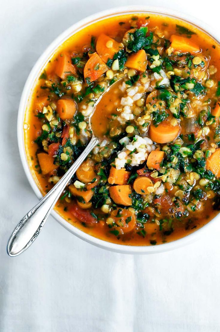 A bowl of carrot red lentil soup with spinach and rice, with a spoon.
