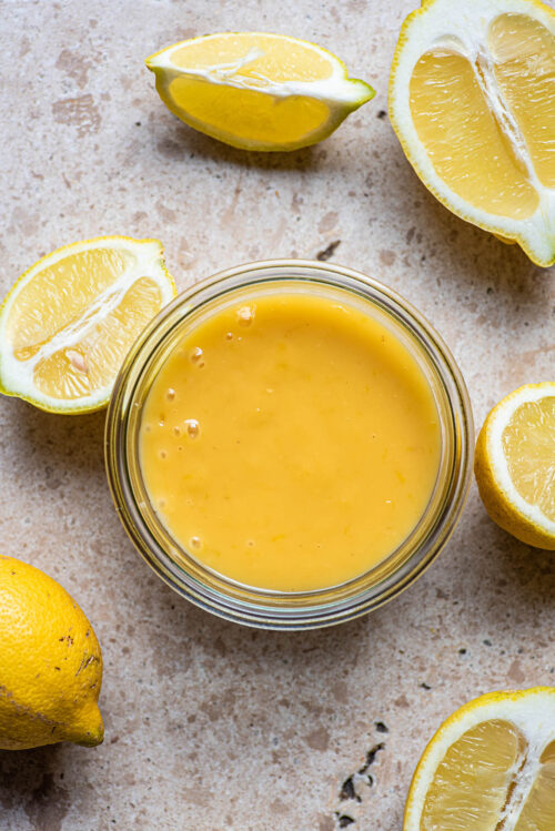 Lemon curd in a glass jar surrounded by lemons.