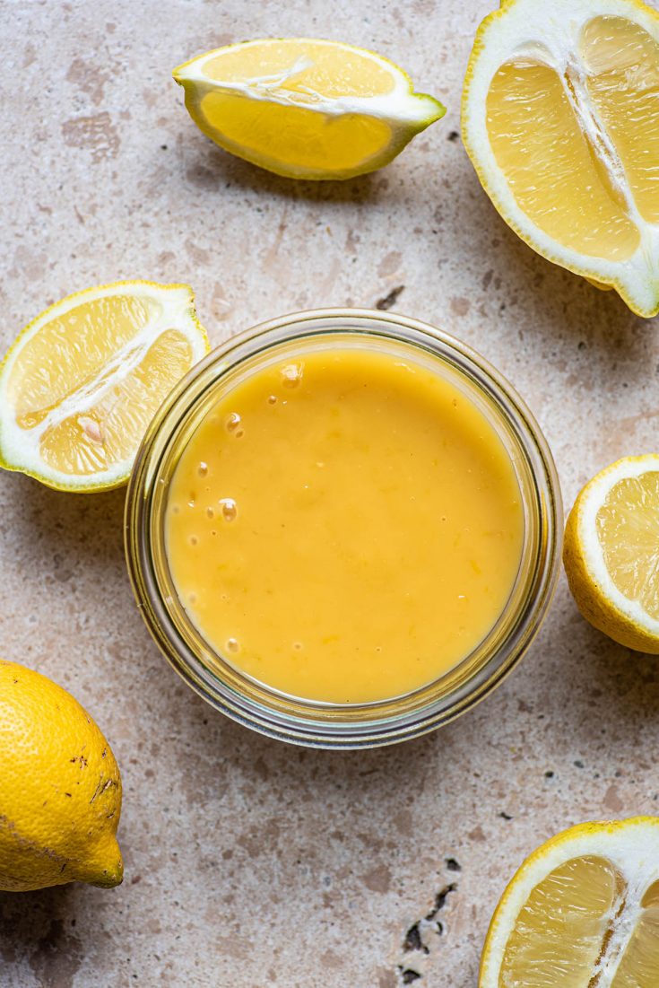 Lemon curd in a glass jar surrounded by lemons.