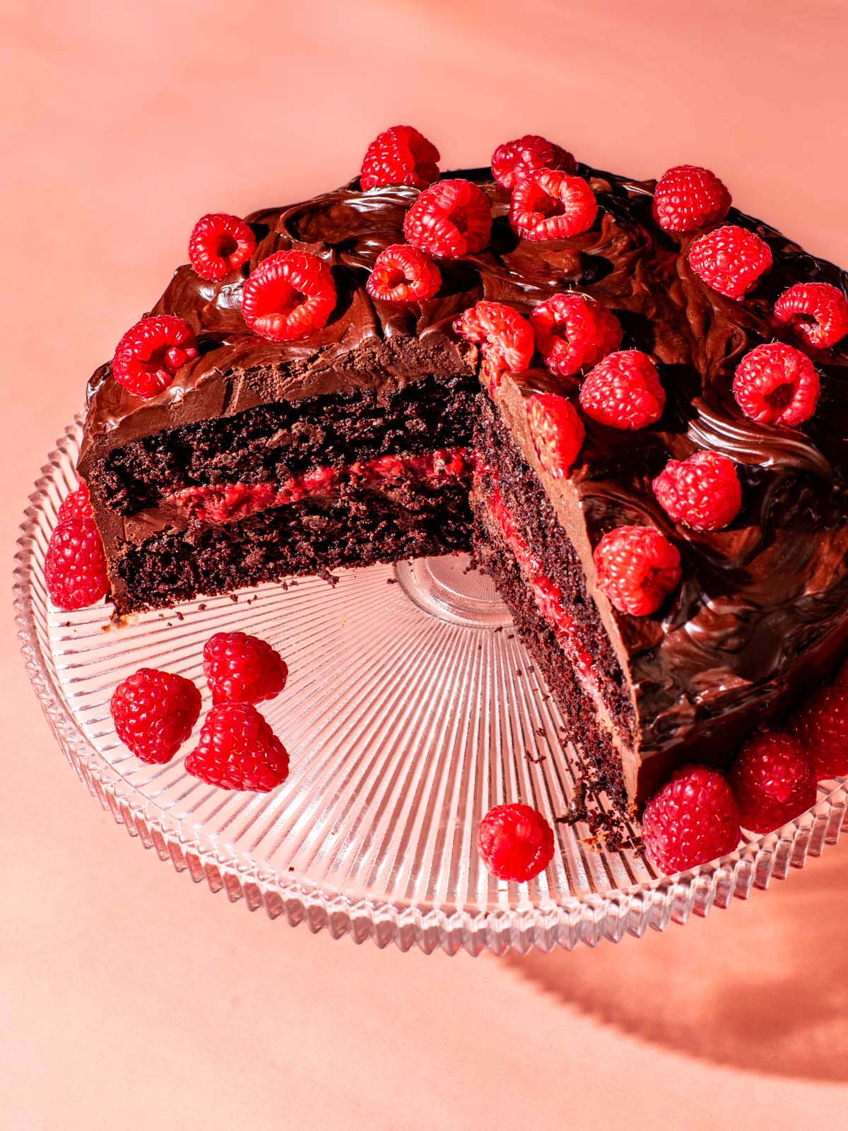 Layered chocolate raspberry cake on a glass cake stand with a couple of slices removed.