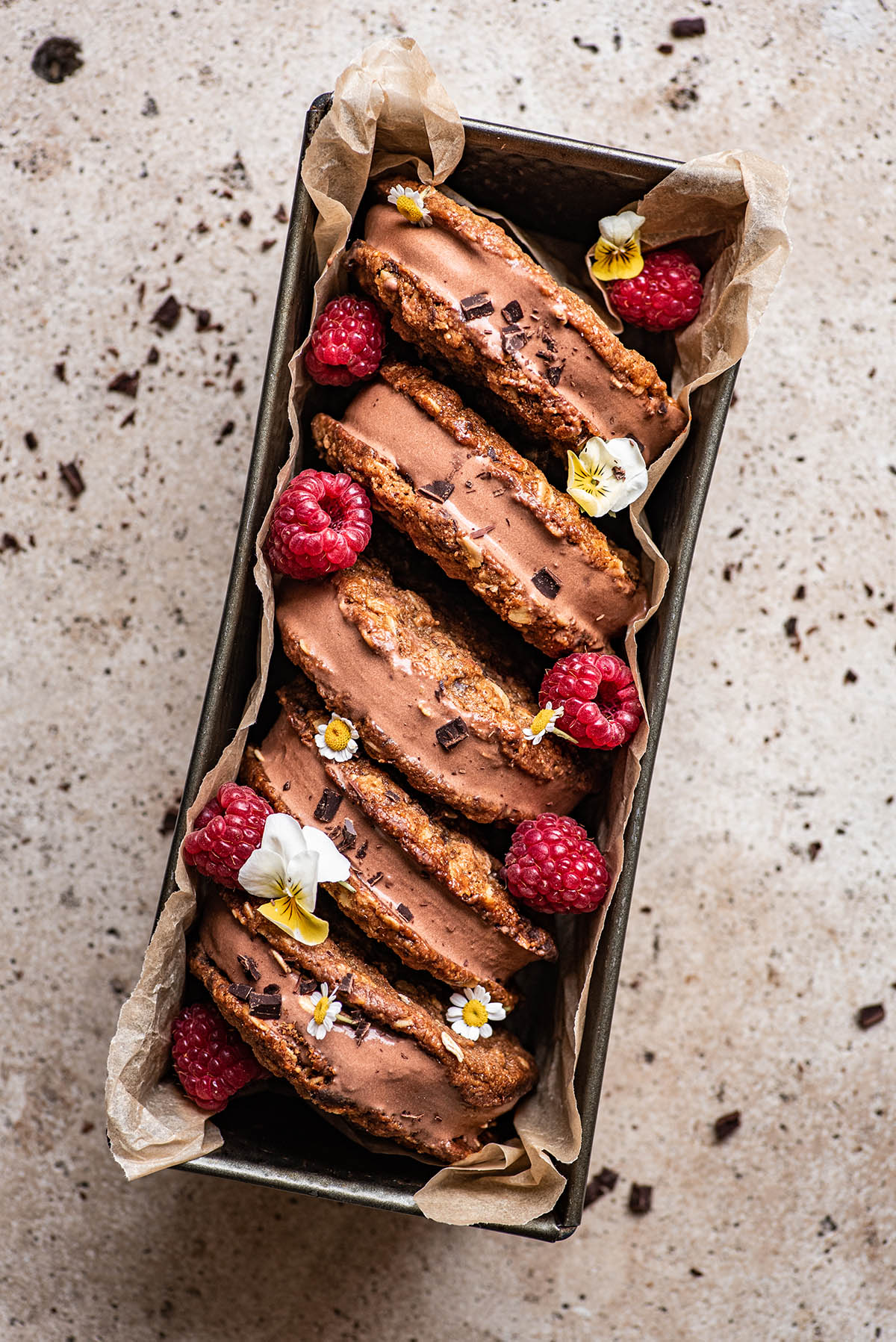Five chocolate ice cream sandwiches lined up in a bread tin, decorated with raspberries and flowers.