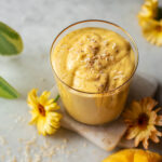 A glass of mango lassi topped with coconut and cardamom, with calendula flowers and mango around it.