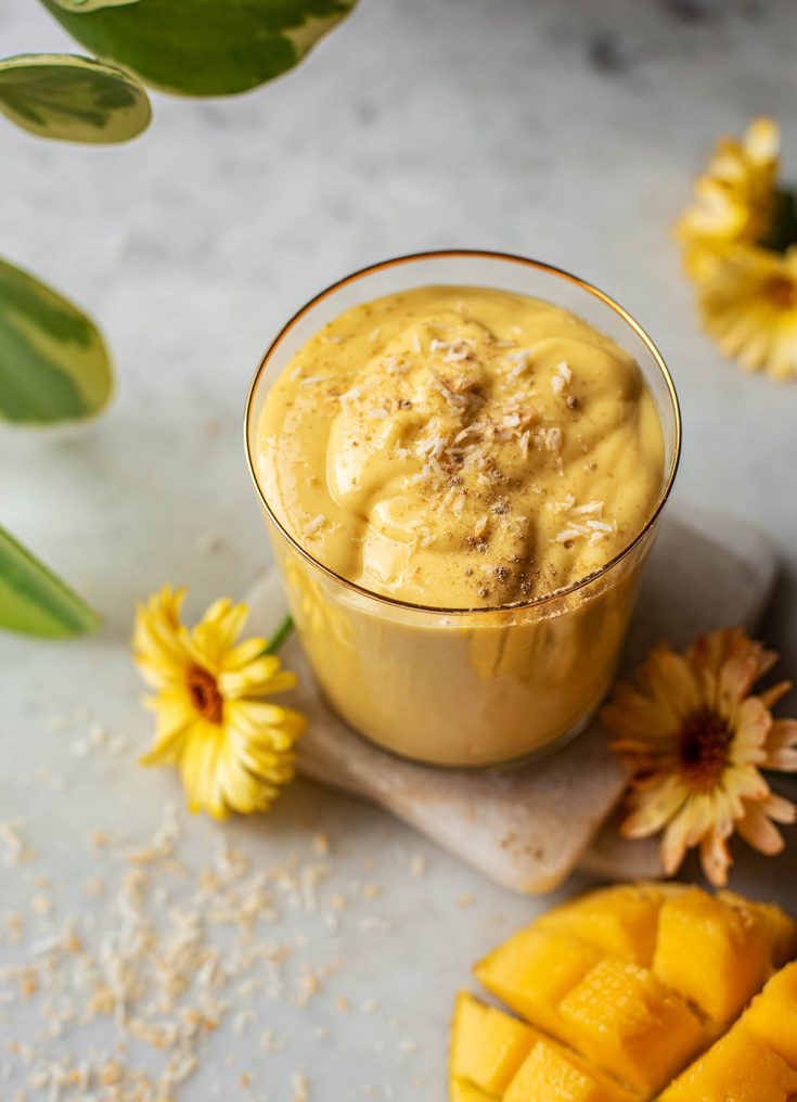 A glass of mango lassi topped with coconut and cardamom, with calendula flowers and mango around it.