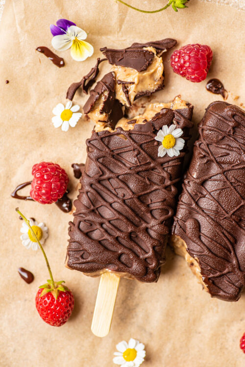 Dark chocolate coated popsicles on parchment paper with flowers and berries, one cut to show interior.