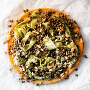 Socca pancake topped with shaved cucumber and lentils on parchment paper.