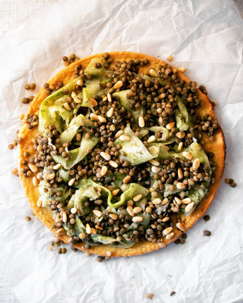 Socca pancake topped with shaved cucumber and lentils on parchment paper.