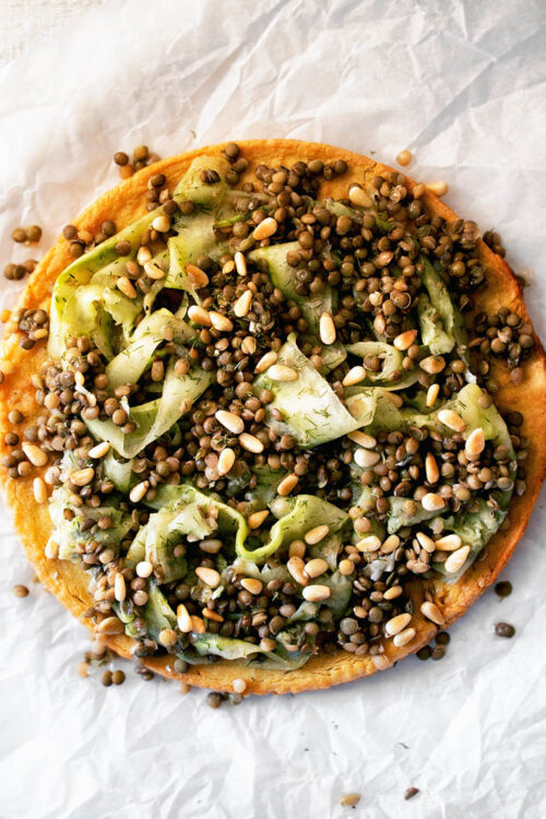 Socca pancake topped with shaved cucumber and lentils on parchment paper.