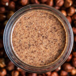 A glass jar of hazelnut butter sitting in a pile of hazelnuts.