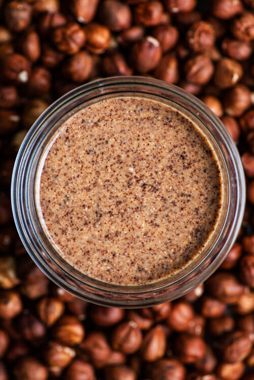 A glass jar of hazelnut butter sitting in a pile of hazelnuts.