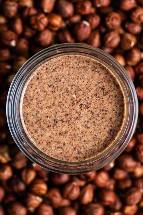 A glass jar of hazelnut butter in a bed of hazelnuts.