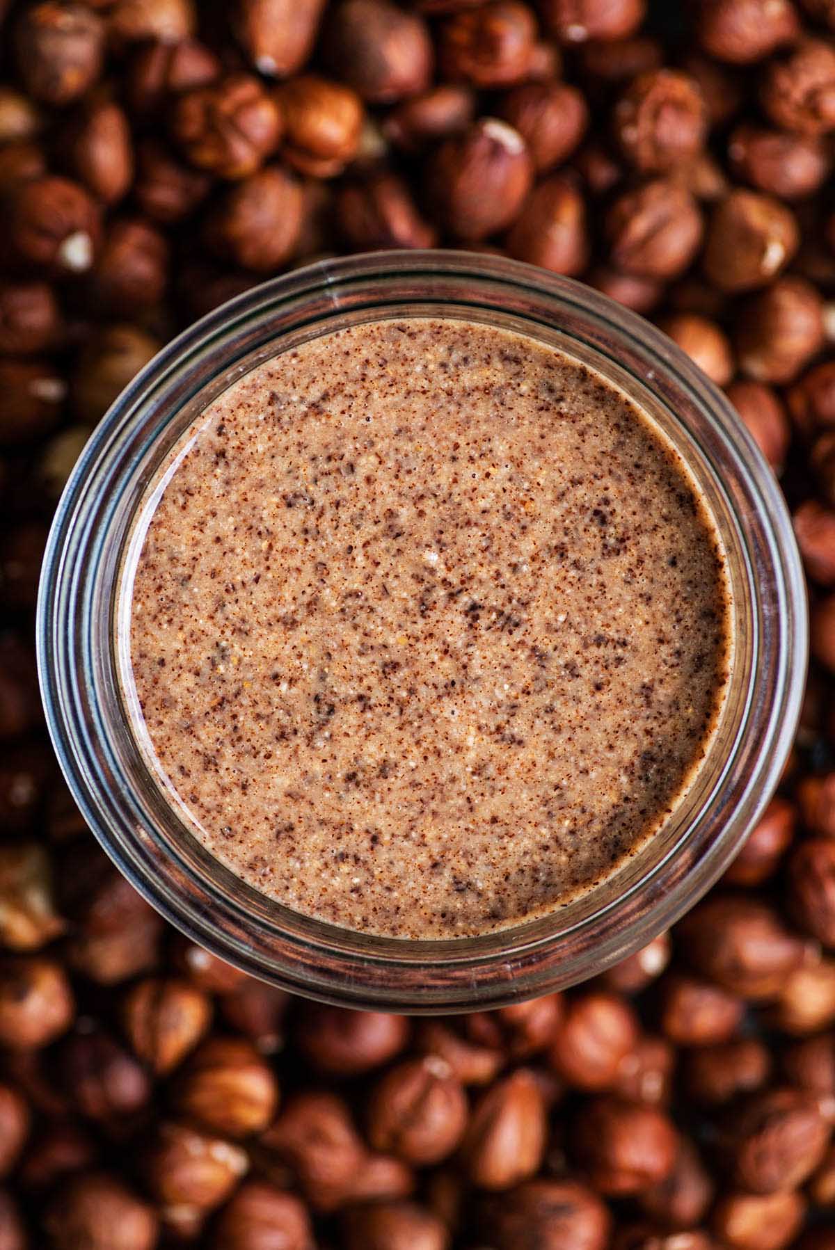 A glass jar of hazelnut butter in a bed of hazelnuts.