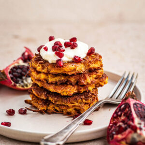 A stack of several sweet potato latkes topped with yogurt and pomegranate seeds.