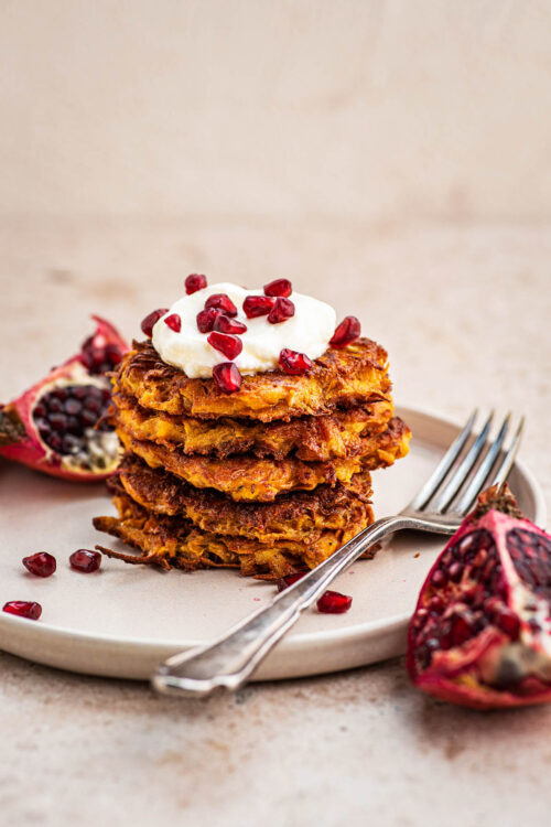 A stack of several sweet potato latkes topped with yogurt and pomegranate seeds.