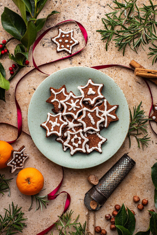 A plate of cookies surrounded with spices, oranges, ribbon, and greenery.