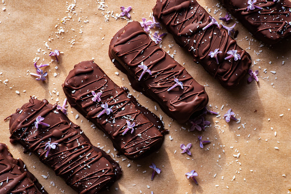 Chocolate coated bars topped with coconut and lilacs in a row, top down.