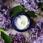 Finished deodorant in a small jar surrounded by lilacs.