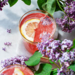 Two glasses of lilac lemonade with ice cubes, lemon slices, and lilacs.