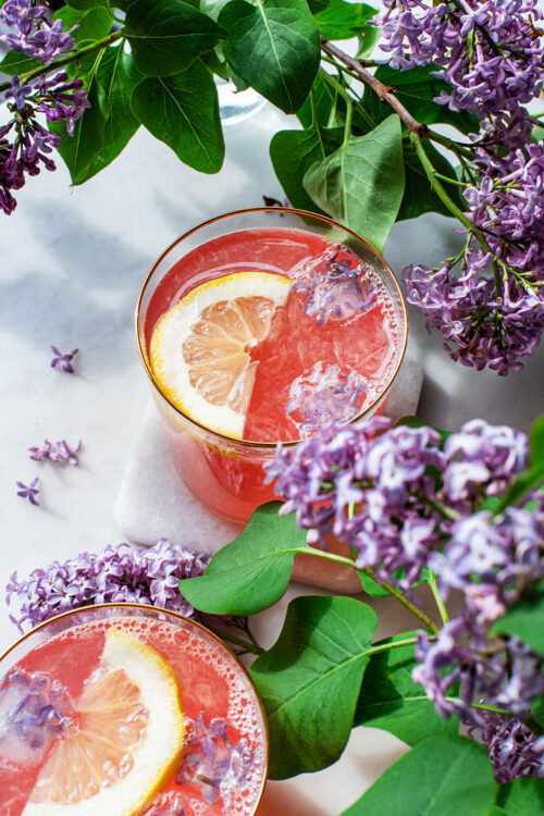 Two glasses of lilac lemonade with ice cubes, lemon slices, and lilacs.