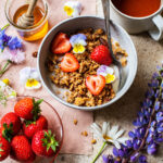 Front angle view of a bowl of granola with strawberries, tea in background and flowers around.