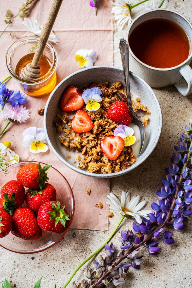 Front angle view of a bowl of granola with strawberries, tea in background and flowers around.