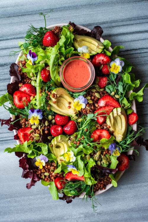 Strawberry salad with edible flowers, avocado, and lentils on a large platter.