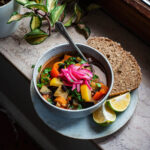 A bowl of soup on a plate with bread and lime pieces.