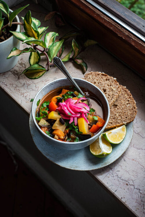 A bowl of soup on a plate with bread and lime pieces.