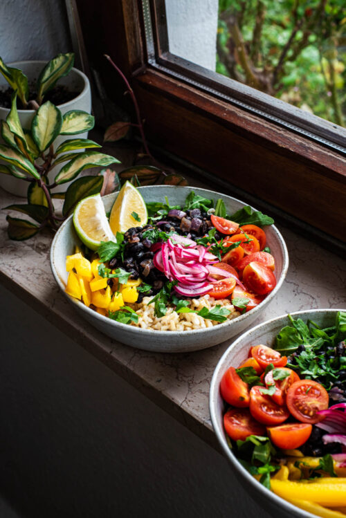 Two burrito bowls on a window ledge with a small plant in background.
