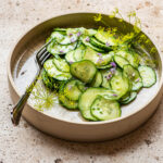 A shallow bowl with cucumber salad, dill flowers, and chive flowers.