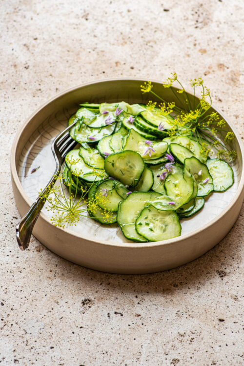 A shallow bowl with cucumber salad, dill flowers, and chive flowers.