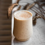 A glass of banana milkshake on a window ledge with dried wheat stalks behind.