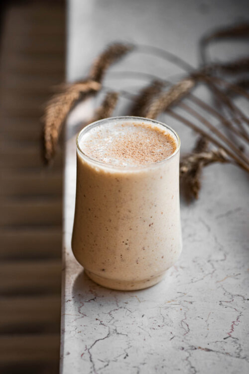 A glass of banana milkshake on a window ledge with dried wheat stalks behind.
